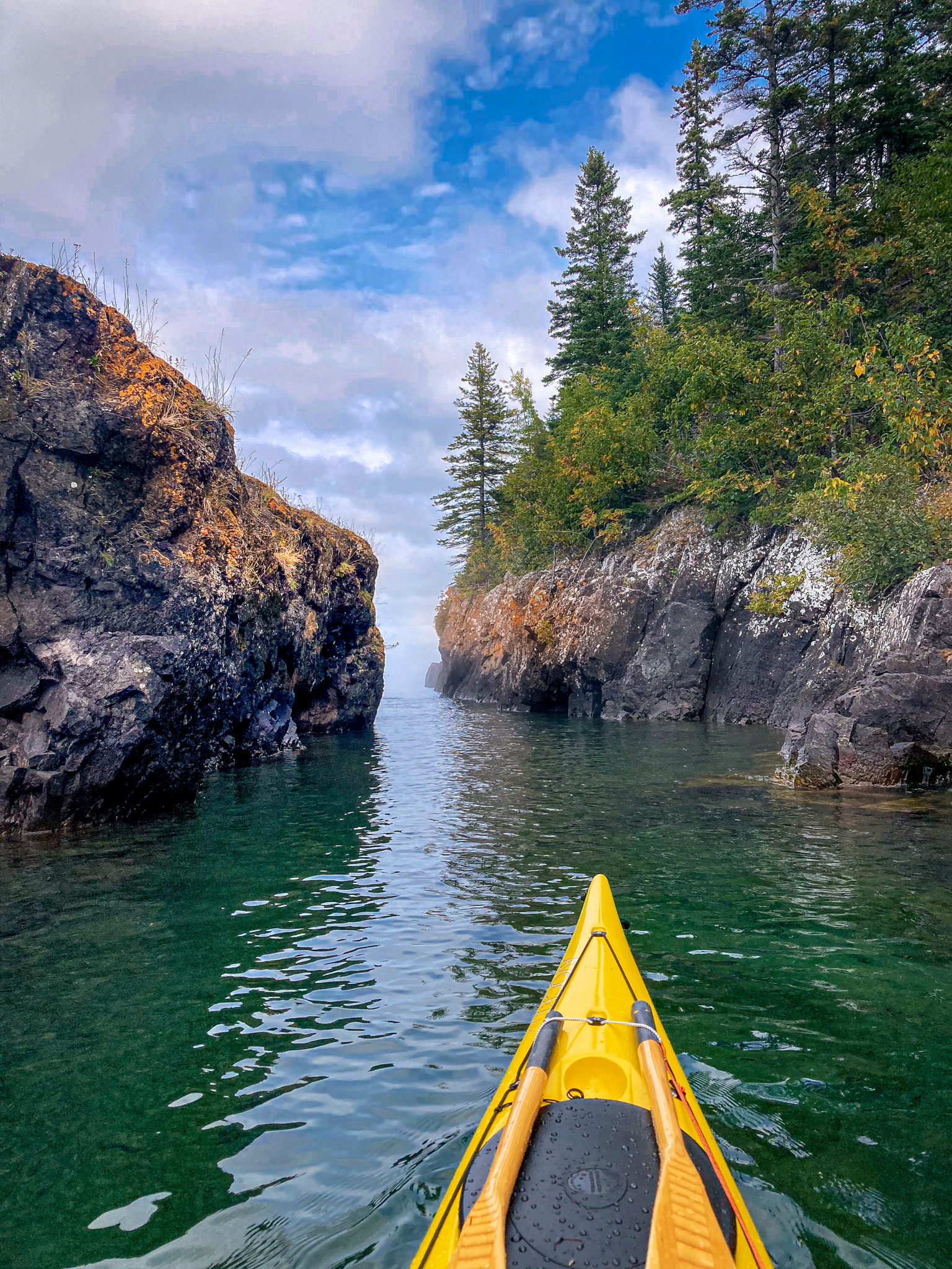Image of the bow of a sea kayak on green water of Lake Superior with a sea stack on the left side of the image and the cliffs of Isle Royale on the right side of the image