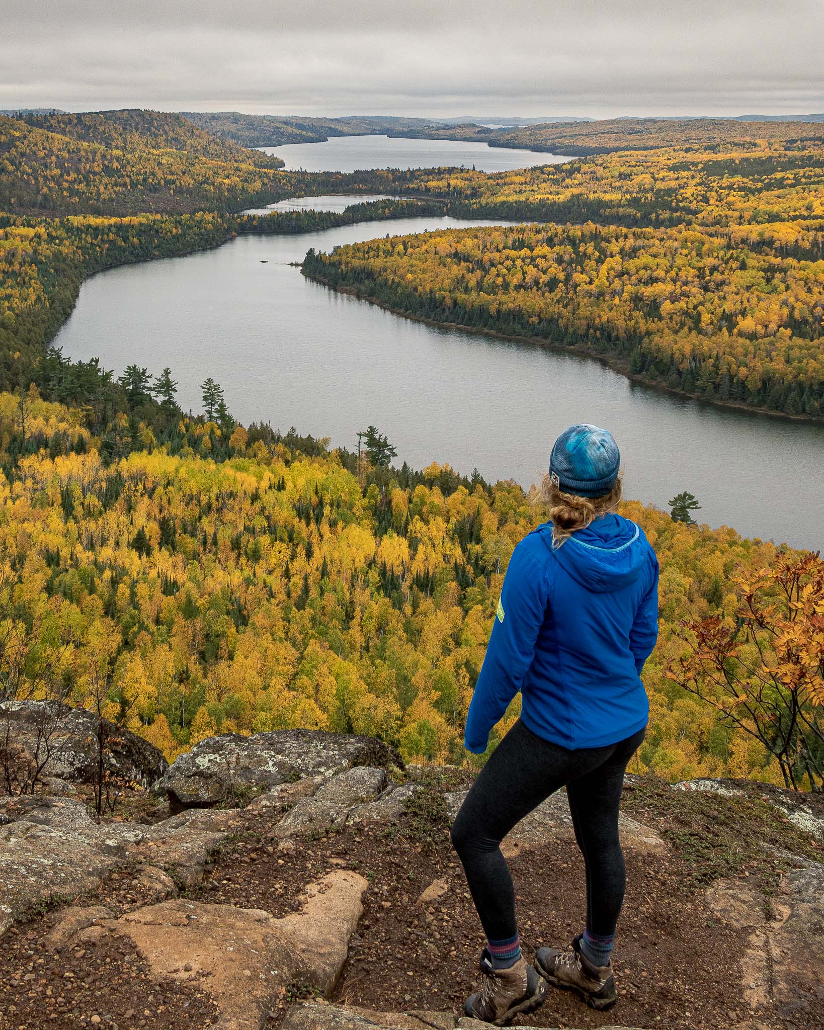 A hiker in blue stands in front of Rose Lake Cliffs Overlook in the BWCA at peak fall color