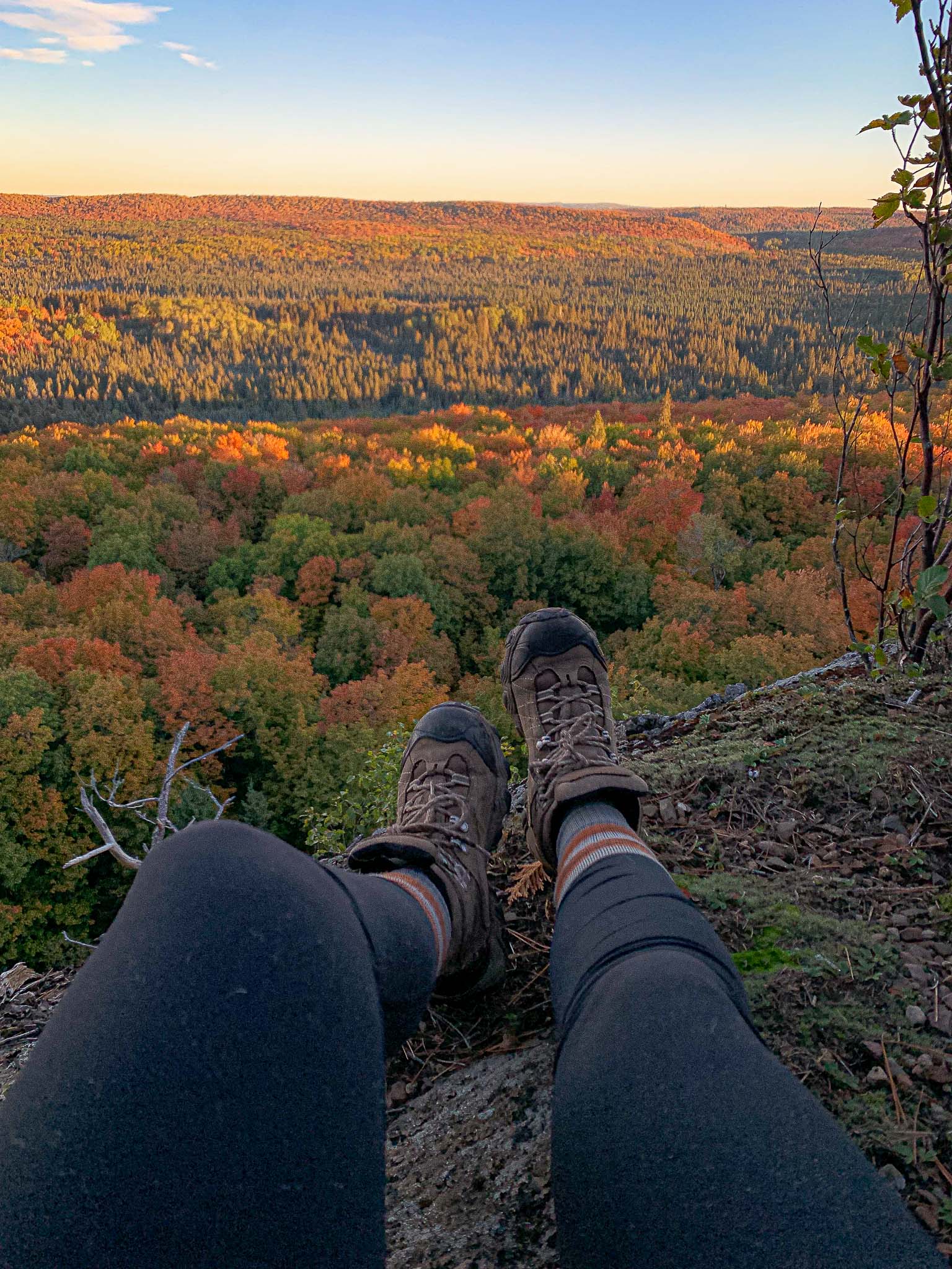 hiking Leveaux Mountain near Oberg Mountain on the North Shore of Lake Superior at Peak Fall Color. A hiker dangles feet off a cliff while early morning sun lights up the valley below.