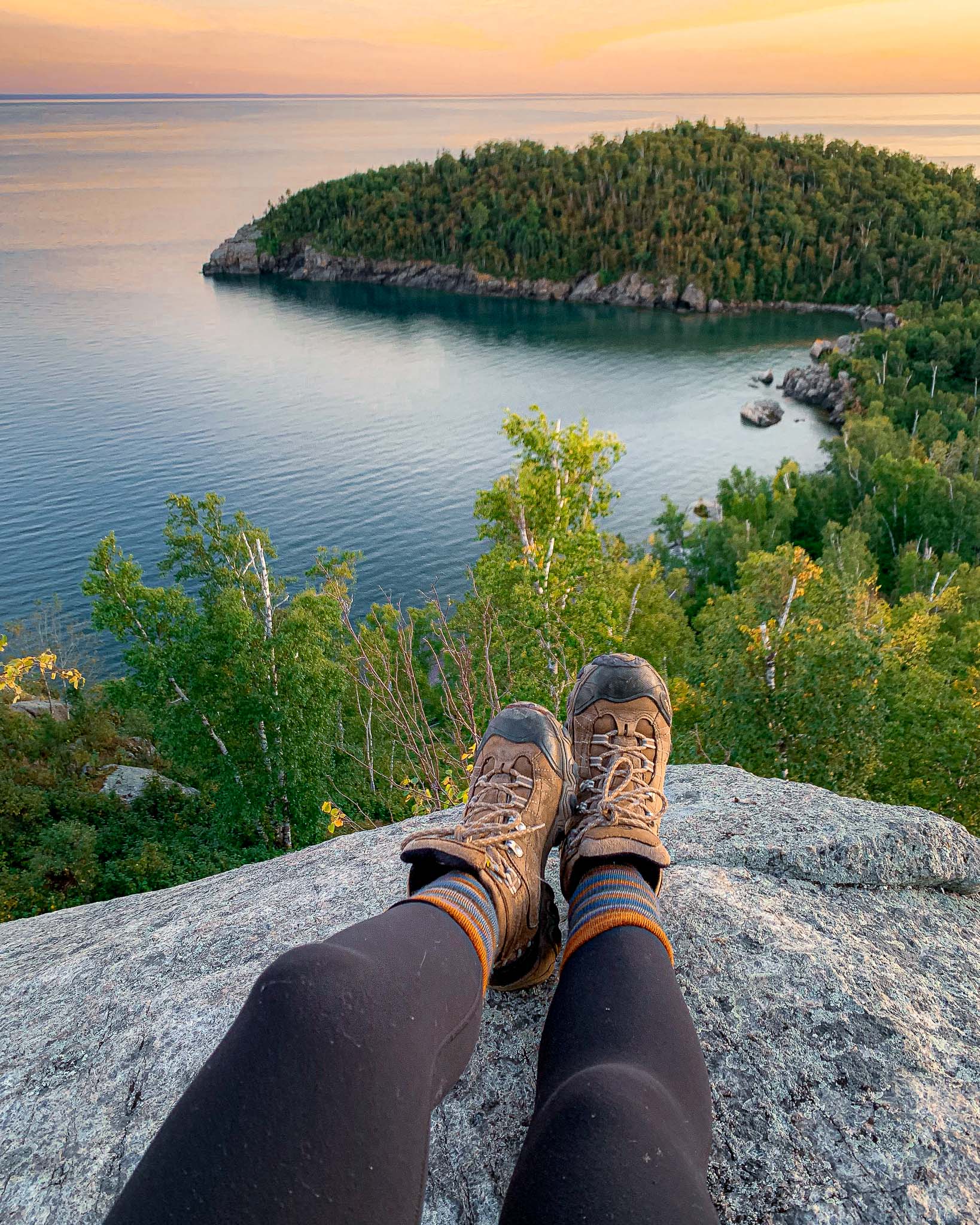 image of Day Hill overlook at Sunset, one of the best hiking trails on Lake Superior, green leaves contrast with blue Lake Superior and a hiker's feet dangle off the rugged cliffs