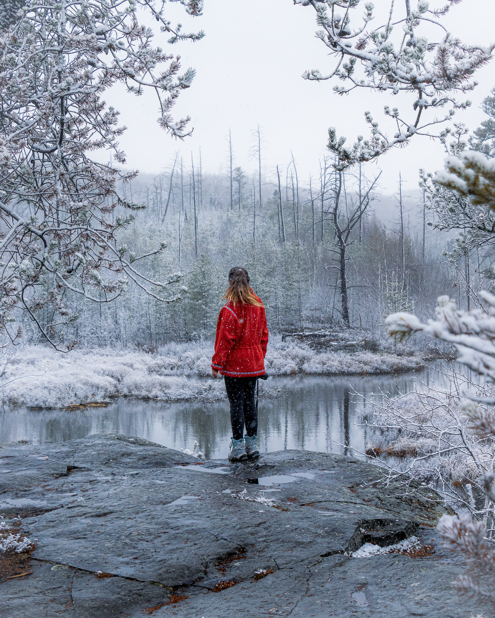 A hiker stands on a ledge overlooking a small pond surrounded by first snow