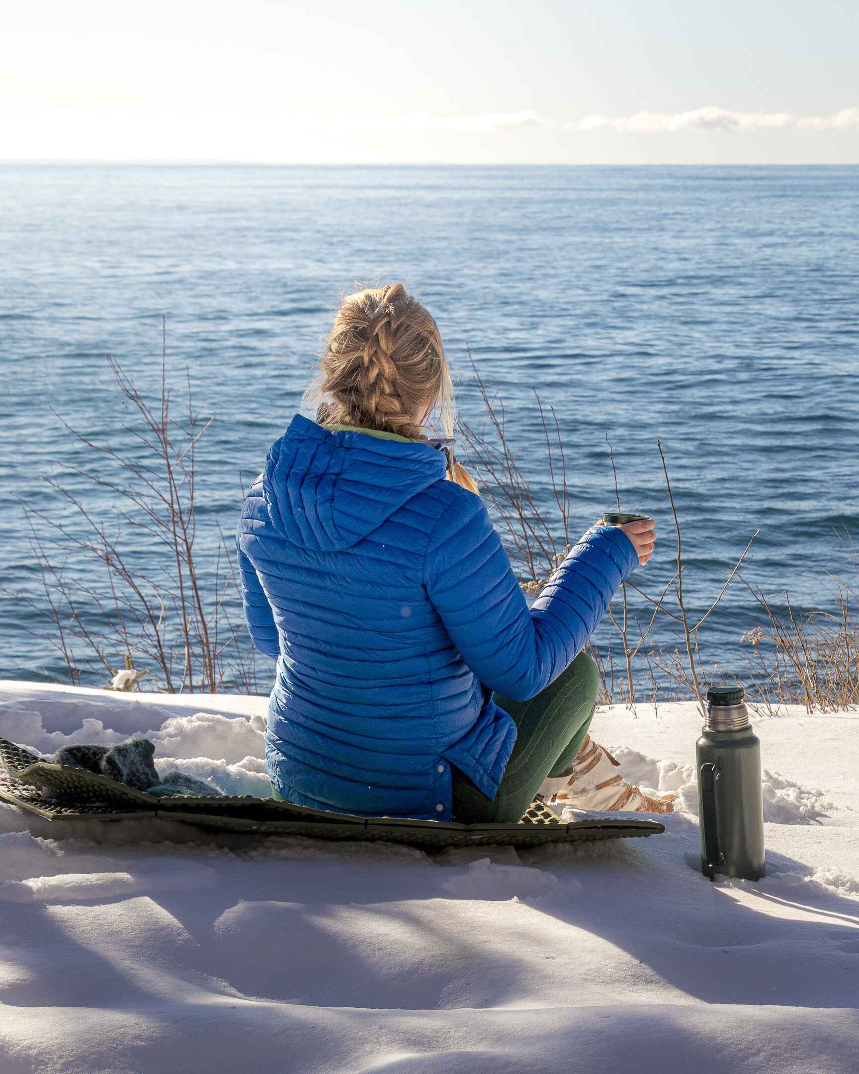 Image of a winter hiker having tea at the edge of a snowy cliff overlooking a lake