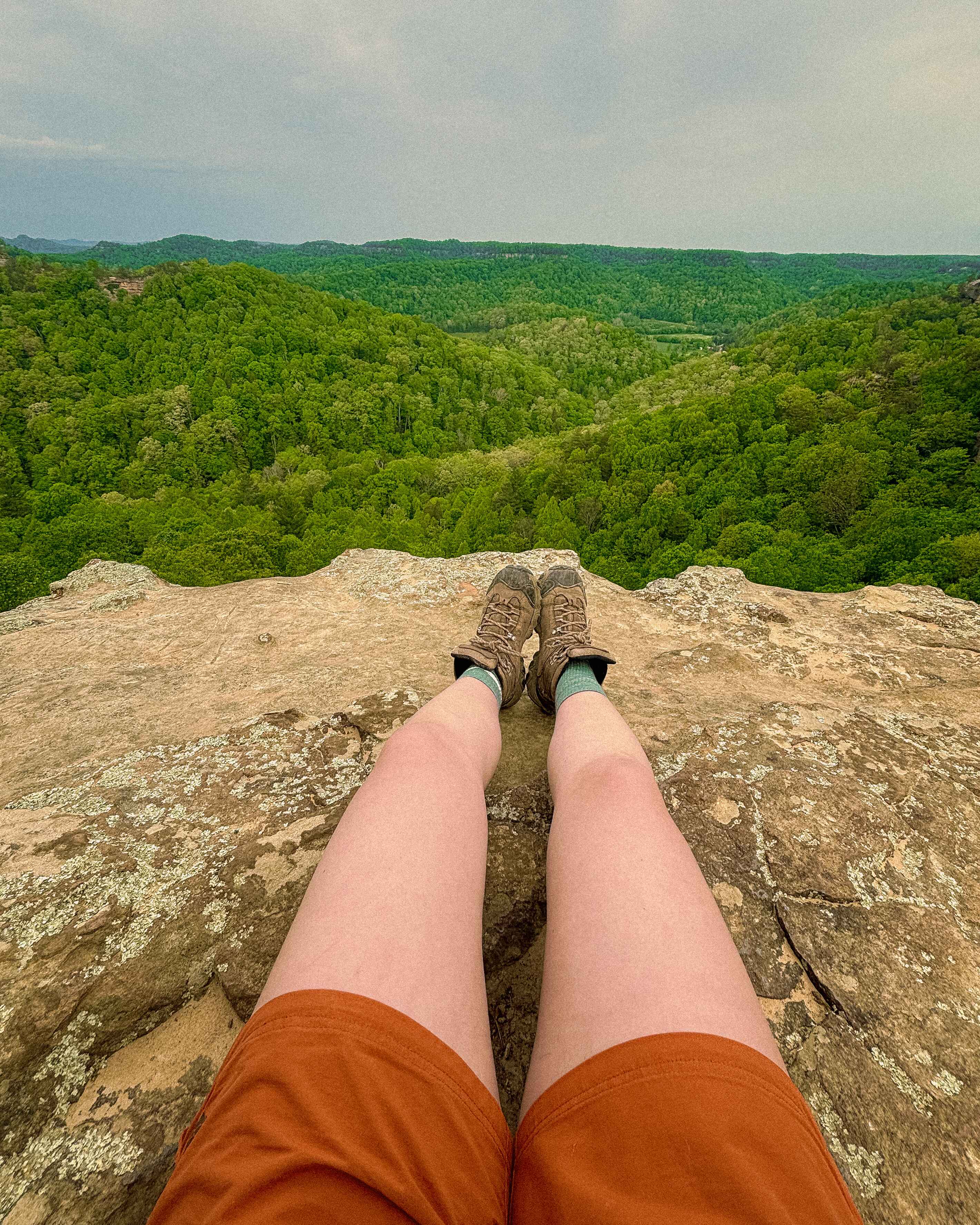 A hikers legs dangle near the edge of a cliff wearing a pair of orange shorts and hiking boots