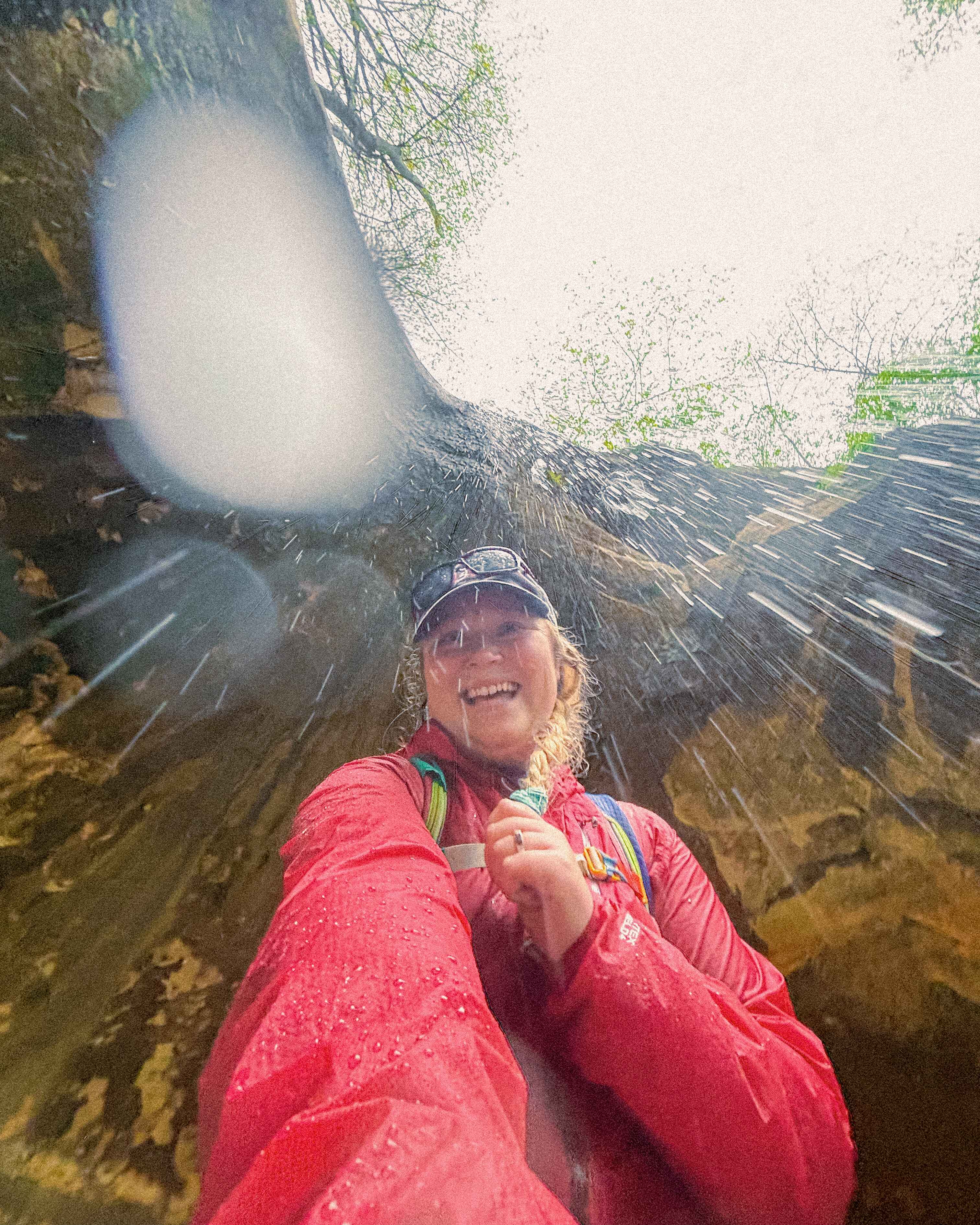 A hiker pauses under a waterfall in Wyalusing State Park, Wisconsin, wearing a raincoat, hat, and backpack