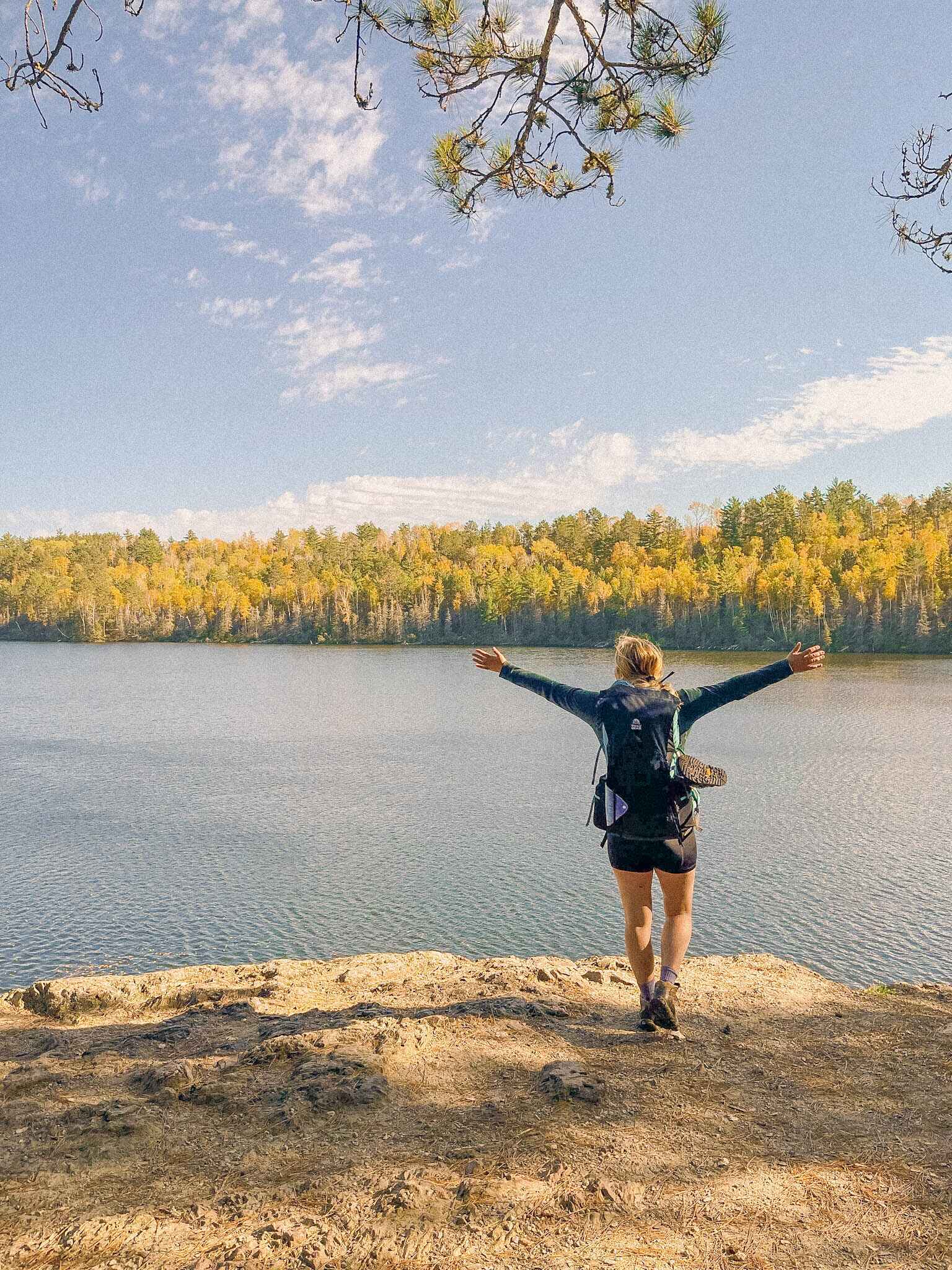 image of a hiker in base layers with her arms up in front of a lake in the fall woods