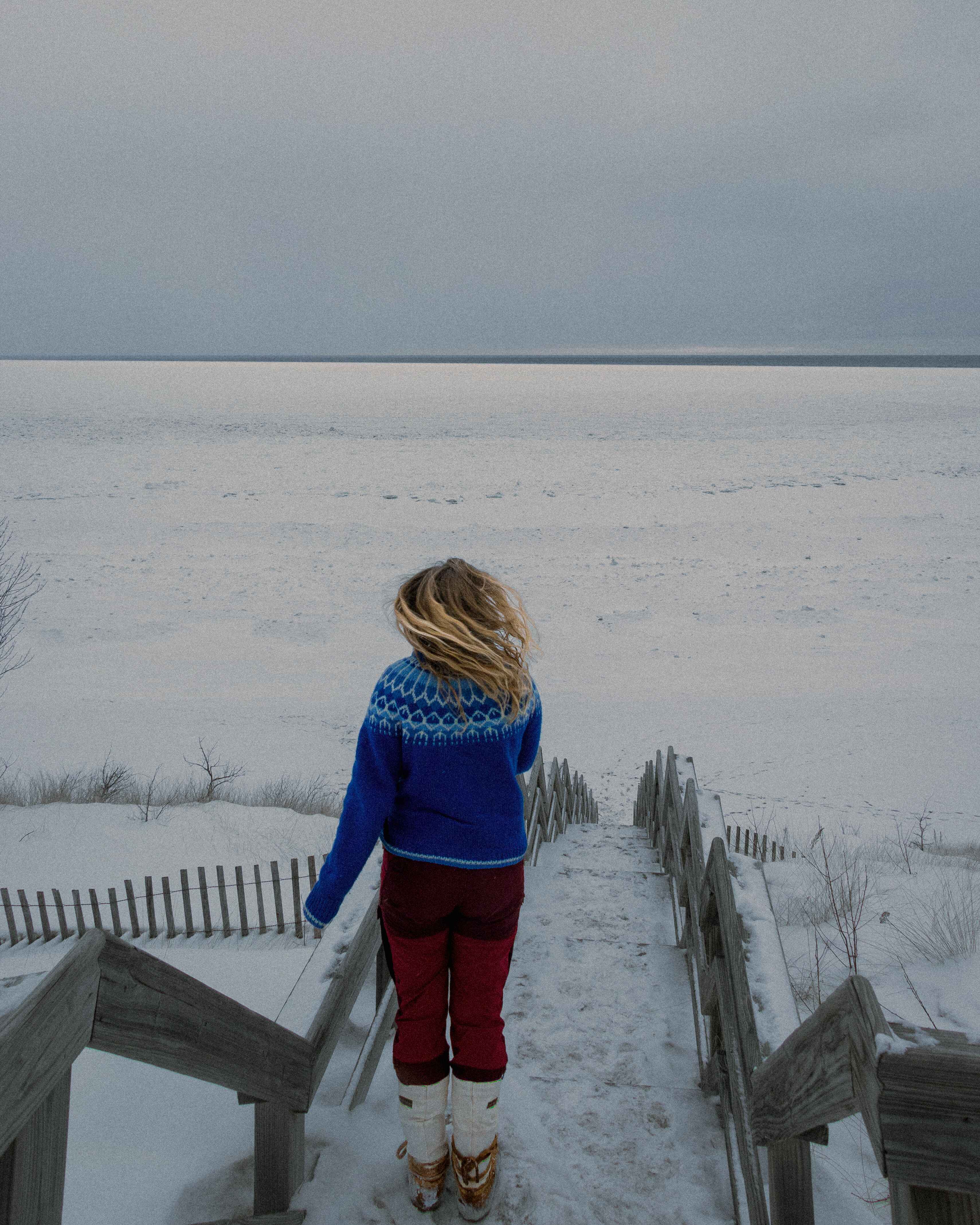 a hiker in a bright sweater stands on a staircase leading down to a frozen lake superior