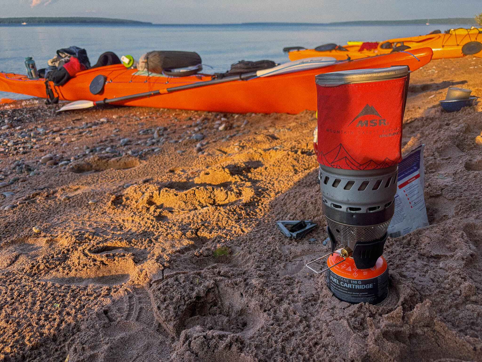 Image of the MSR WindBurner camping stove and a dehydrated meal on a beach with seak kayaks and islands on Lake Superior in the background