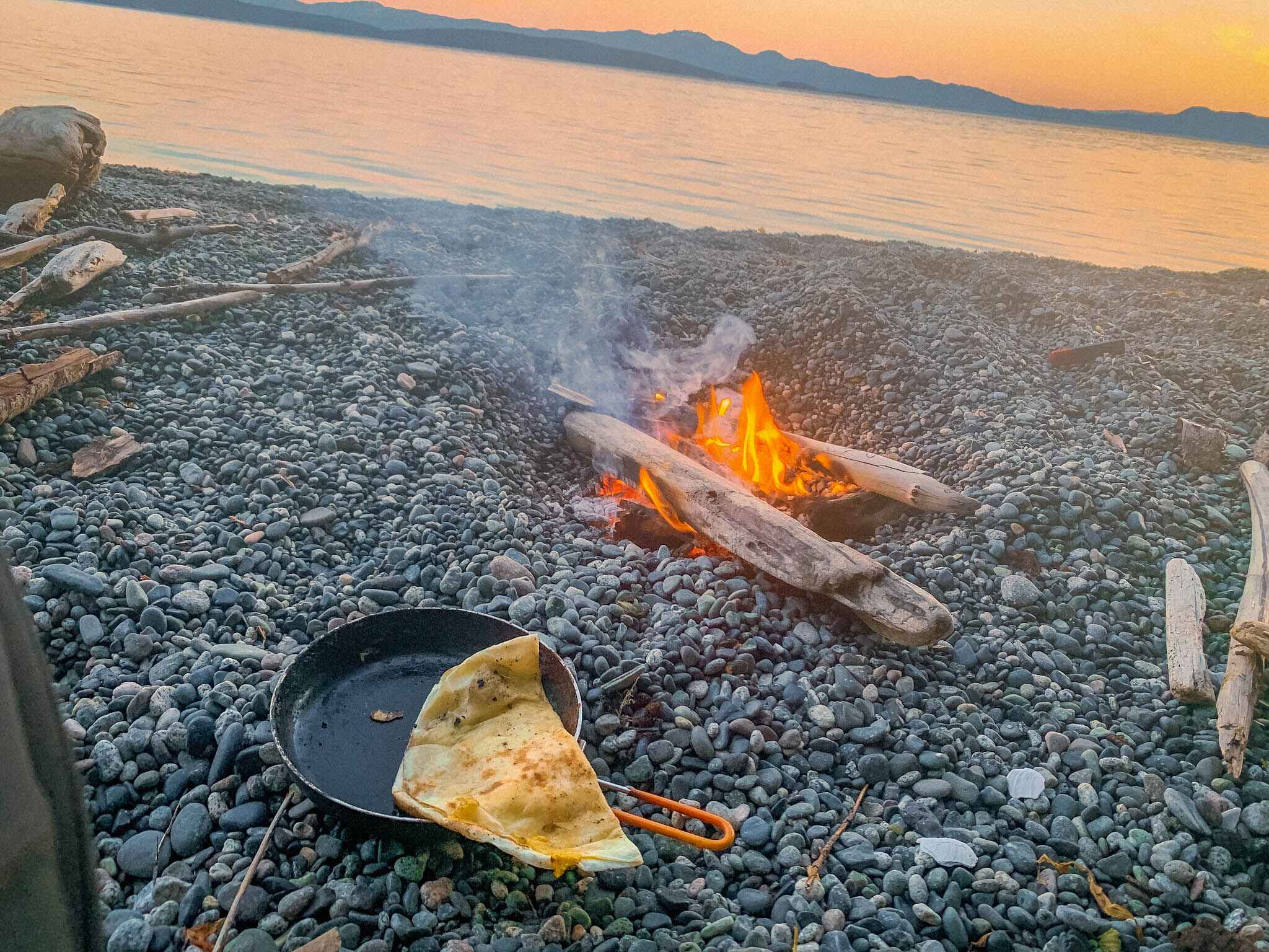 Image of a quesadilla in a camp cooking pan cooked over a small beach fire at sunrise with mountains in the distance on Vancouver Island, British Columbia 