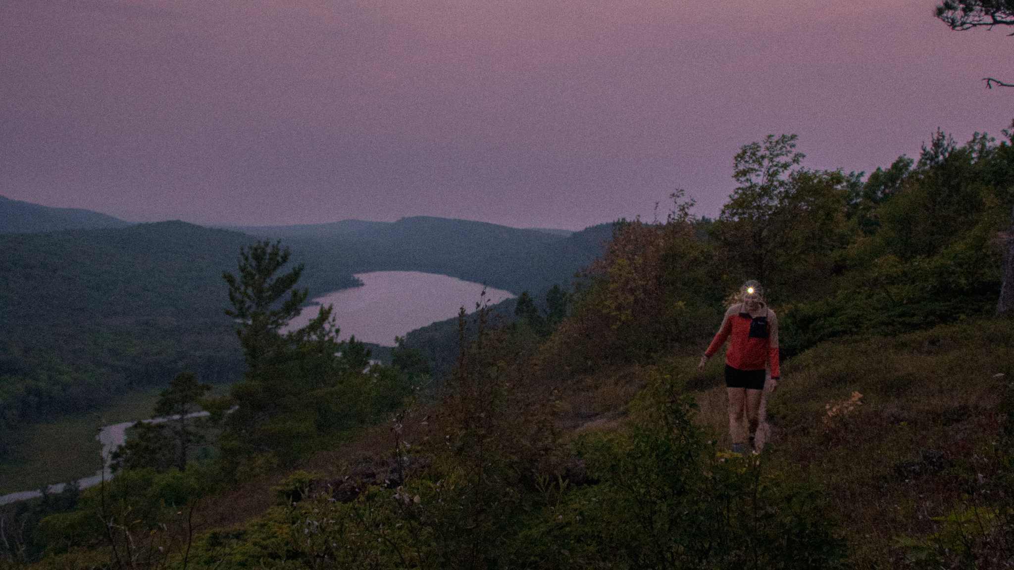 Image of a hiker on a trail above a mountain lake at dusk wearing a headlamp