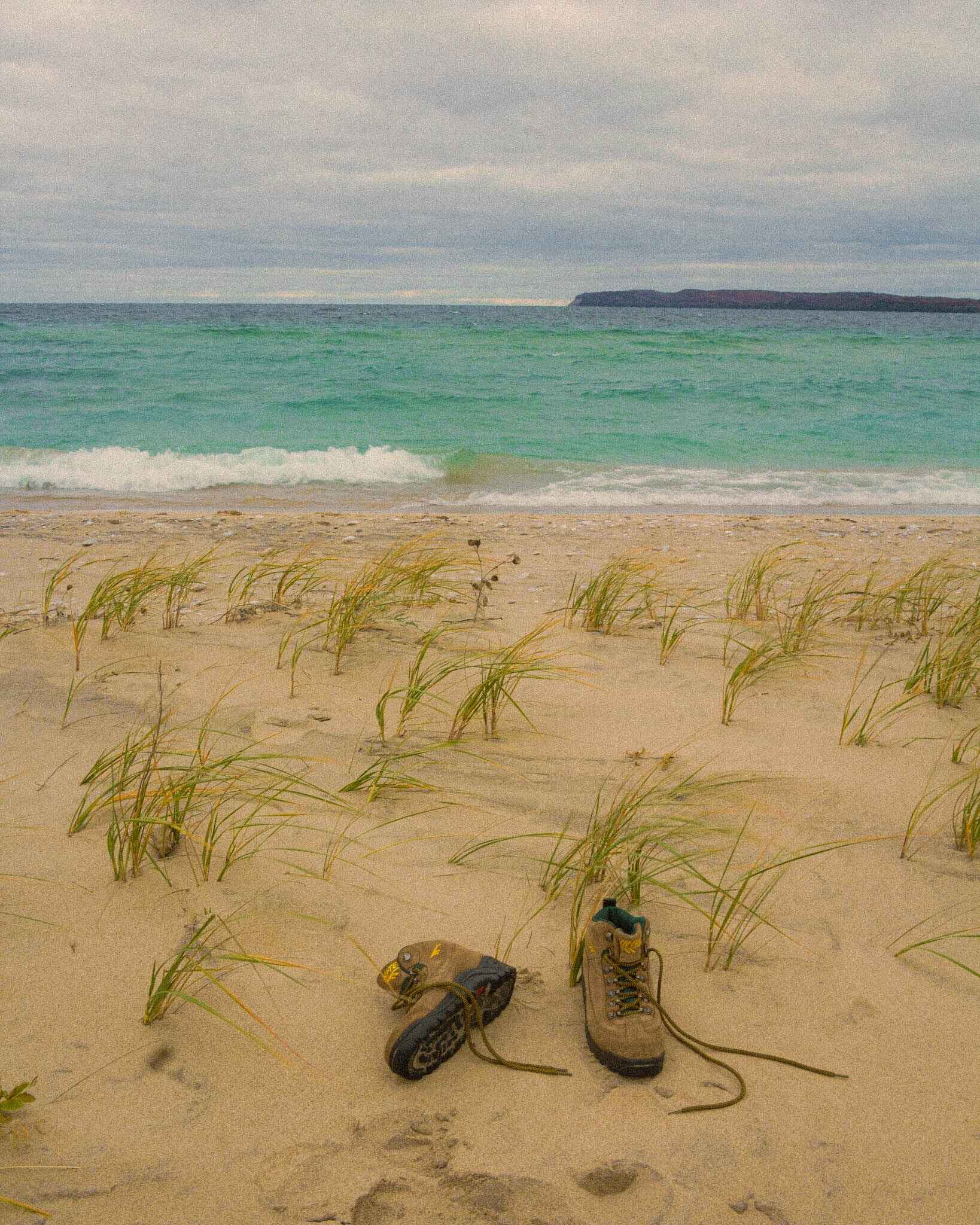 Hiking boots lay in the sand on a beach in the Sleeping Bear Dunes near Traverse City, with blue great water in the middle of the image