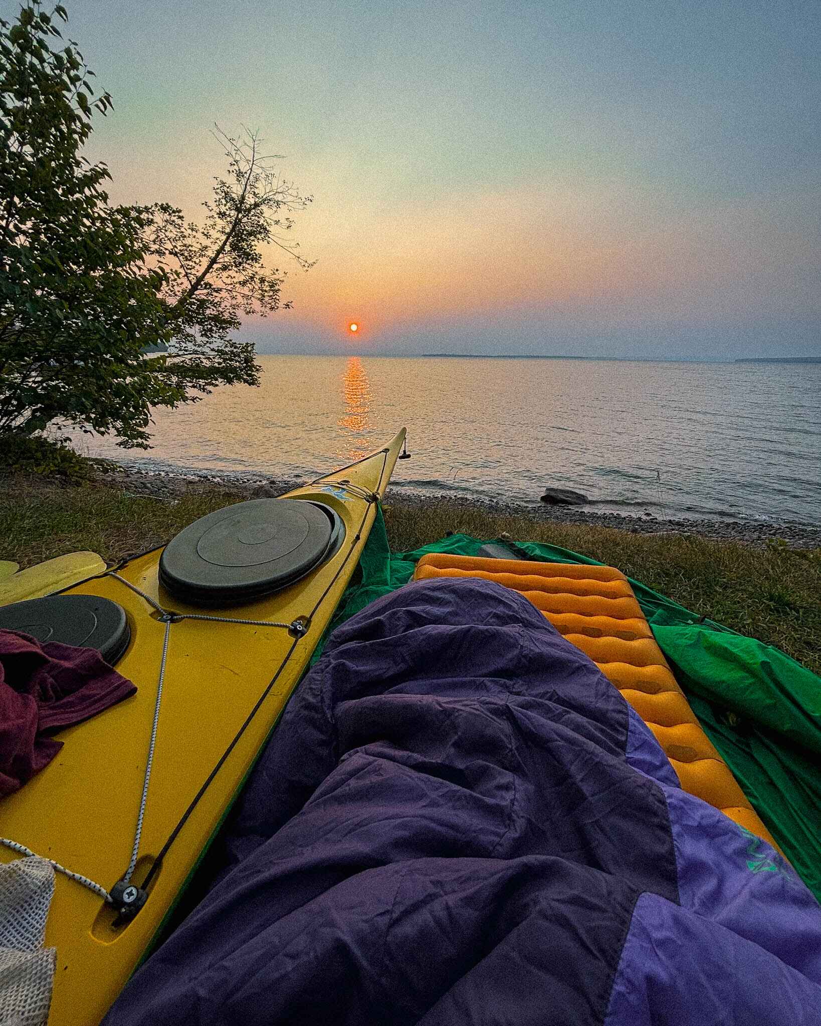 image of nemo tensor sleeping pad on the ground with a sleeping bag on top next to a yellow kayak looking out over the water and a sunrise at a cowboy camp