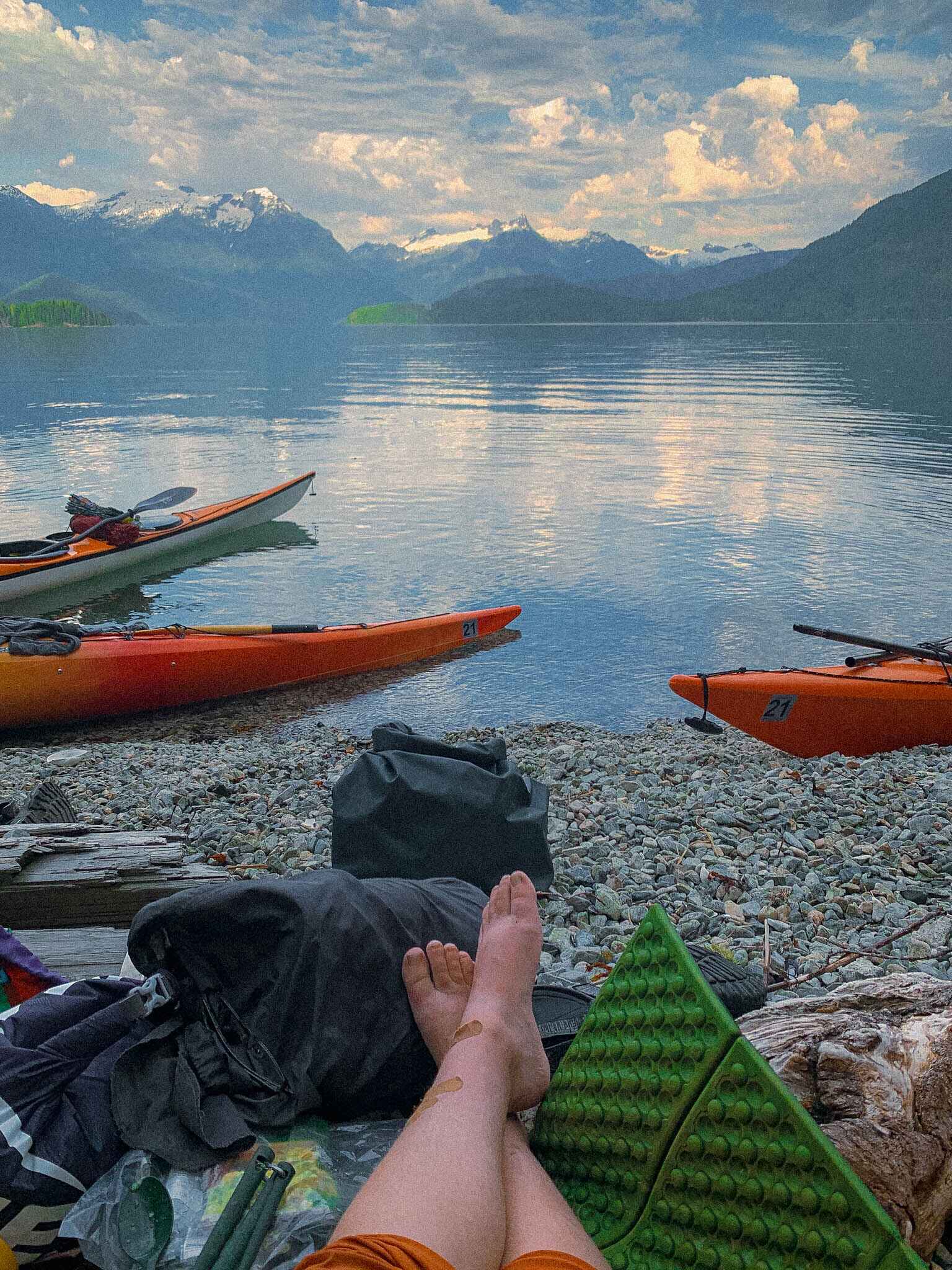 Image of a woman sitting with legs crossed on a sleeping pad looking out over Desolation Sound with kayaks floating in the water and mountains in the background