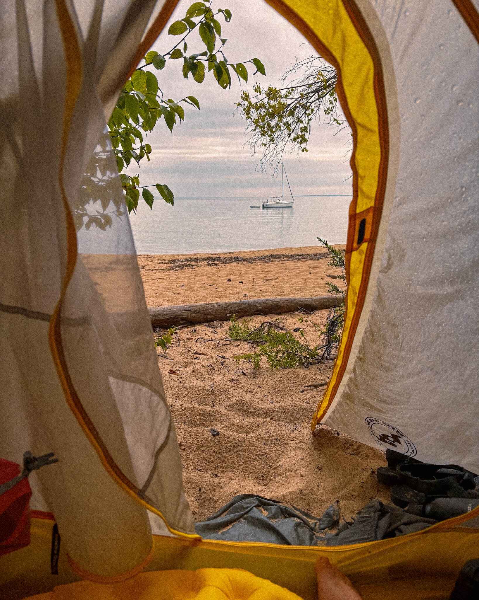 looking out from a tent with a sleeping pad from the woods out at a beach with a sailboat and island in the distance