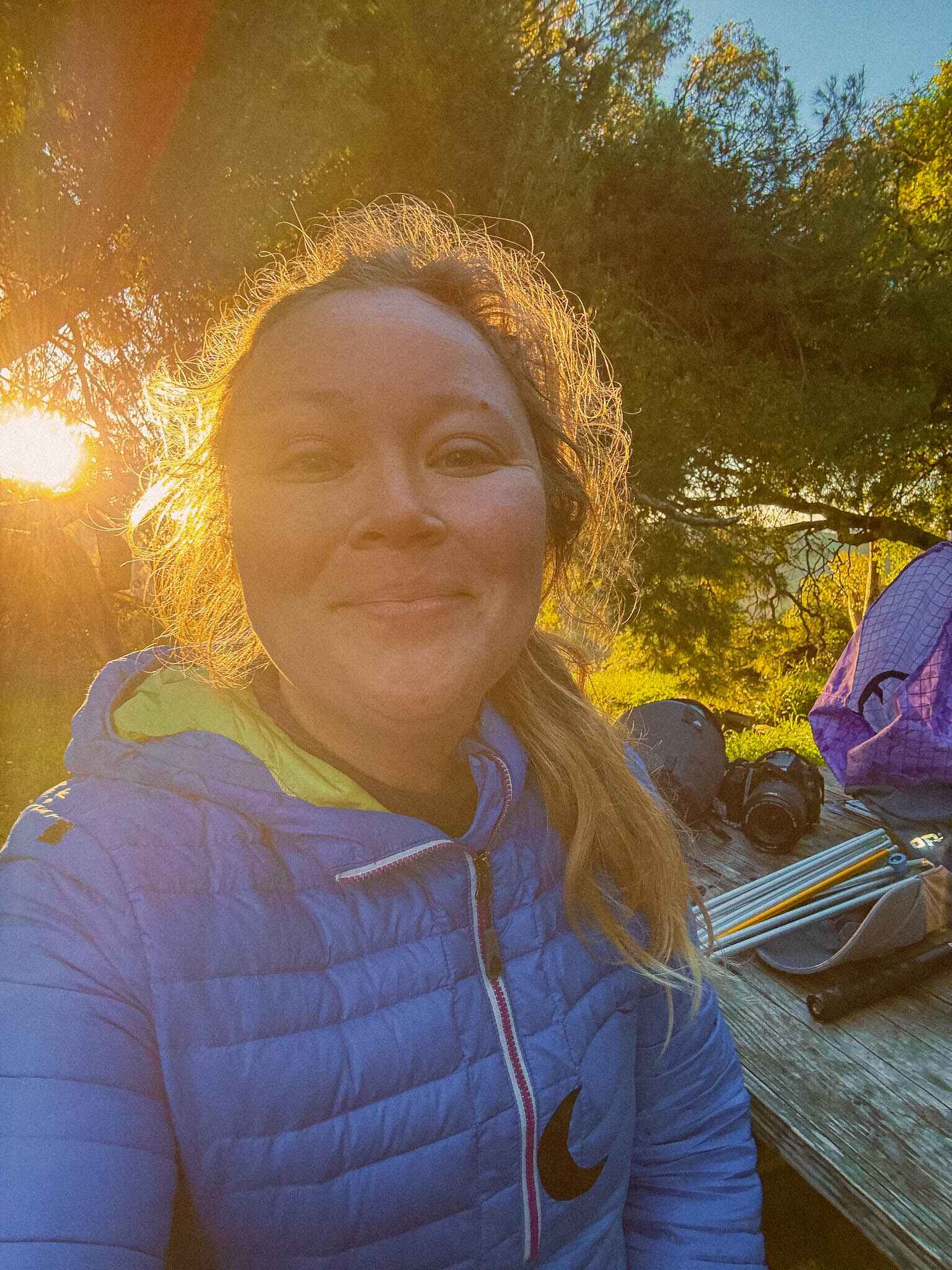 a selfie of the article author (Maddy) while hiking the trail -- a blond woman with frizzy hair wearing a blue jacket sits by a picnic table and smiles at the camera