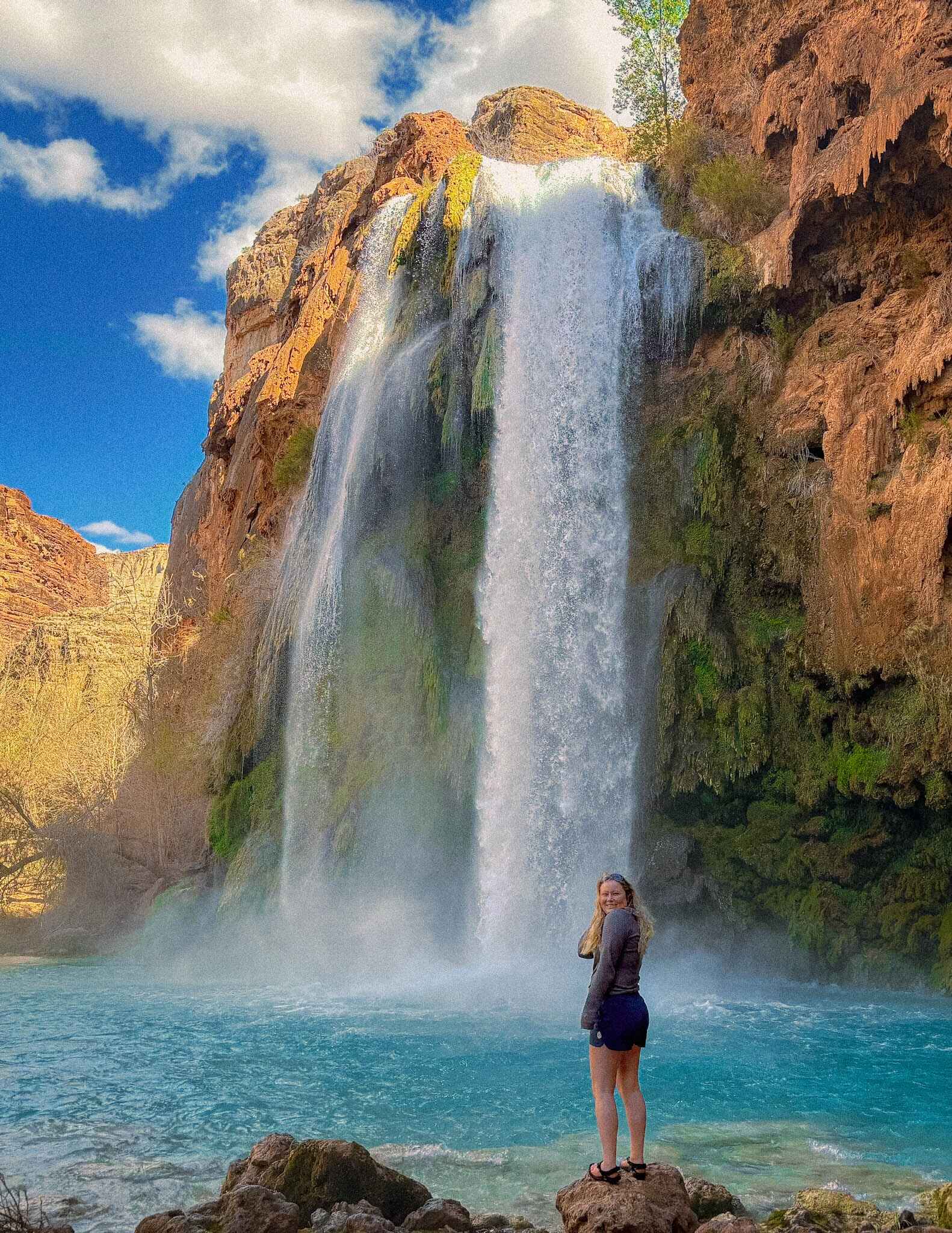 A hiker stands in front of a waterfall in hiking sandals, a wool shirt, and quick dry hiking shorts