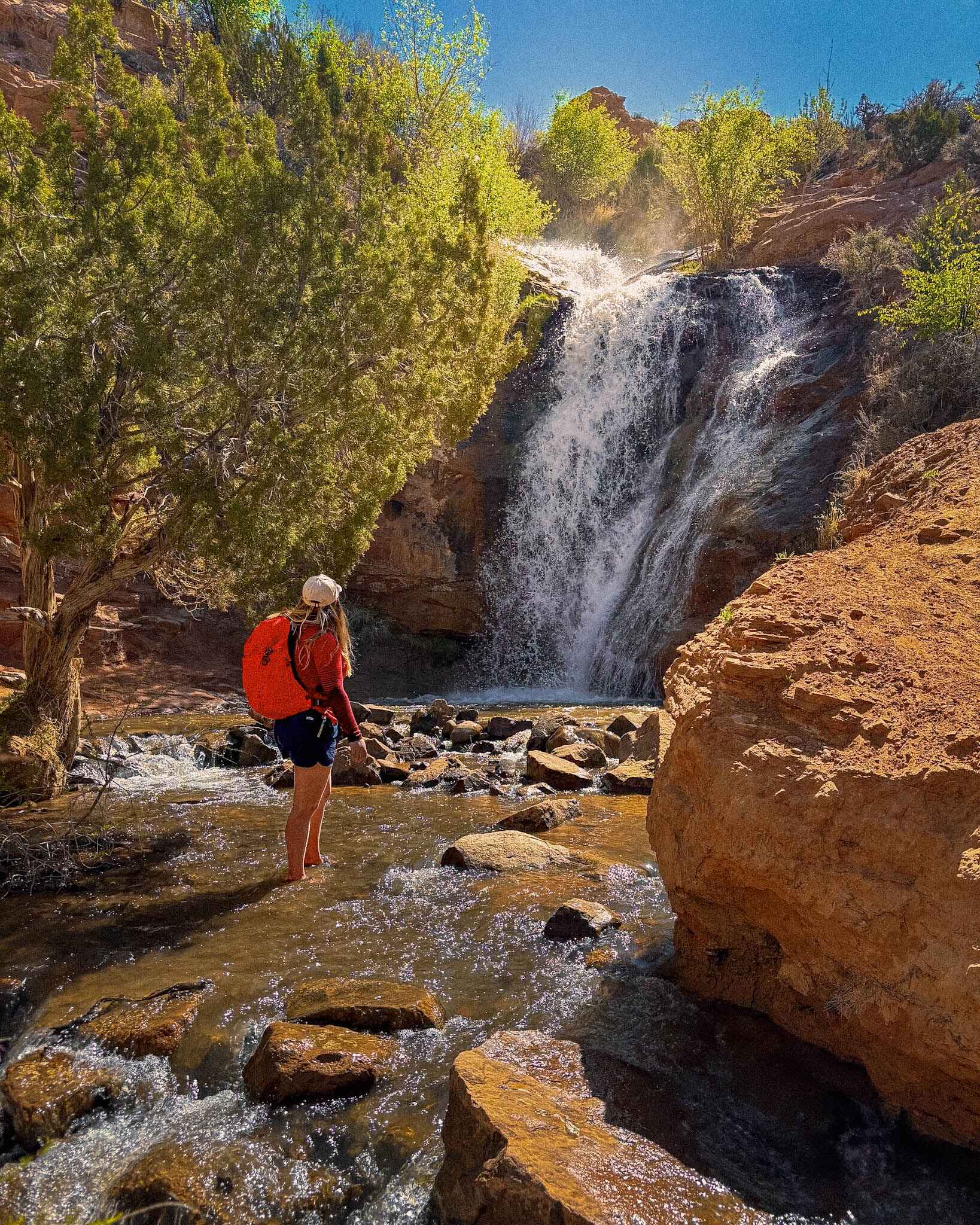 A hiker stands in front of a waterfall in Moab wearing a hiking backpack, sunshirt, and quick dry shorts