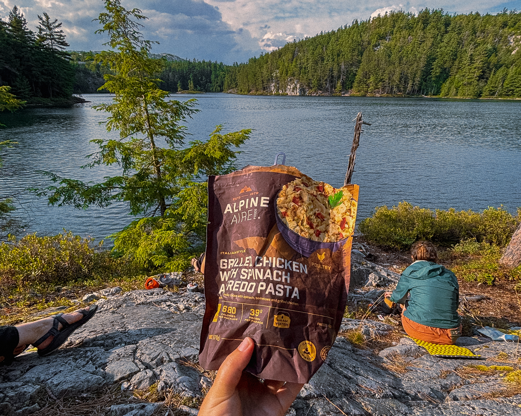 A backpacking meal held up to the camera in front of a rugged Canadian Lake