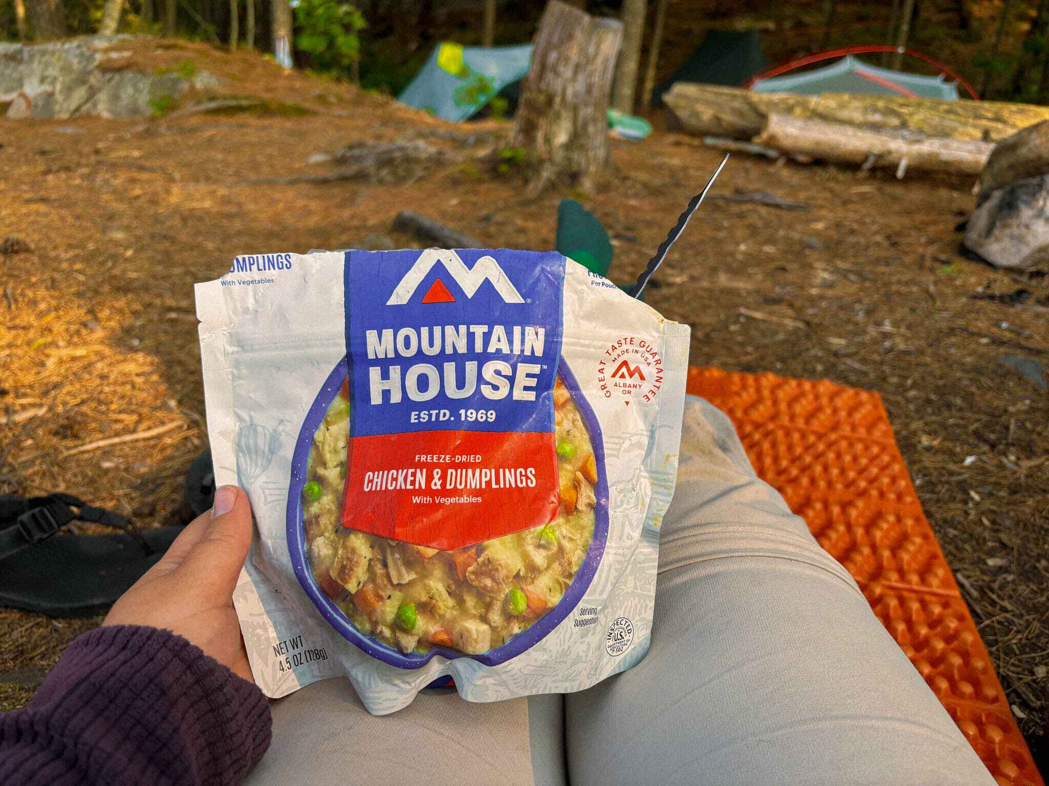 A hiker eats Mountain House Chicken and Dumpling at a campsite with tents in the background