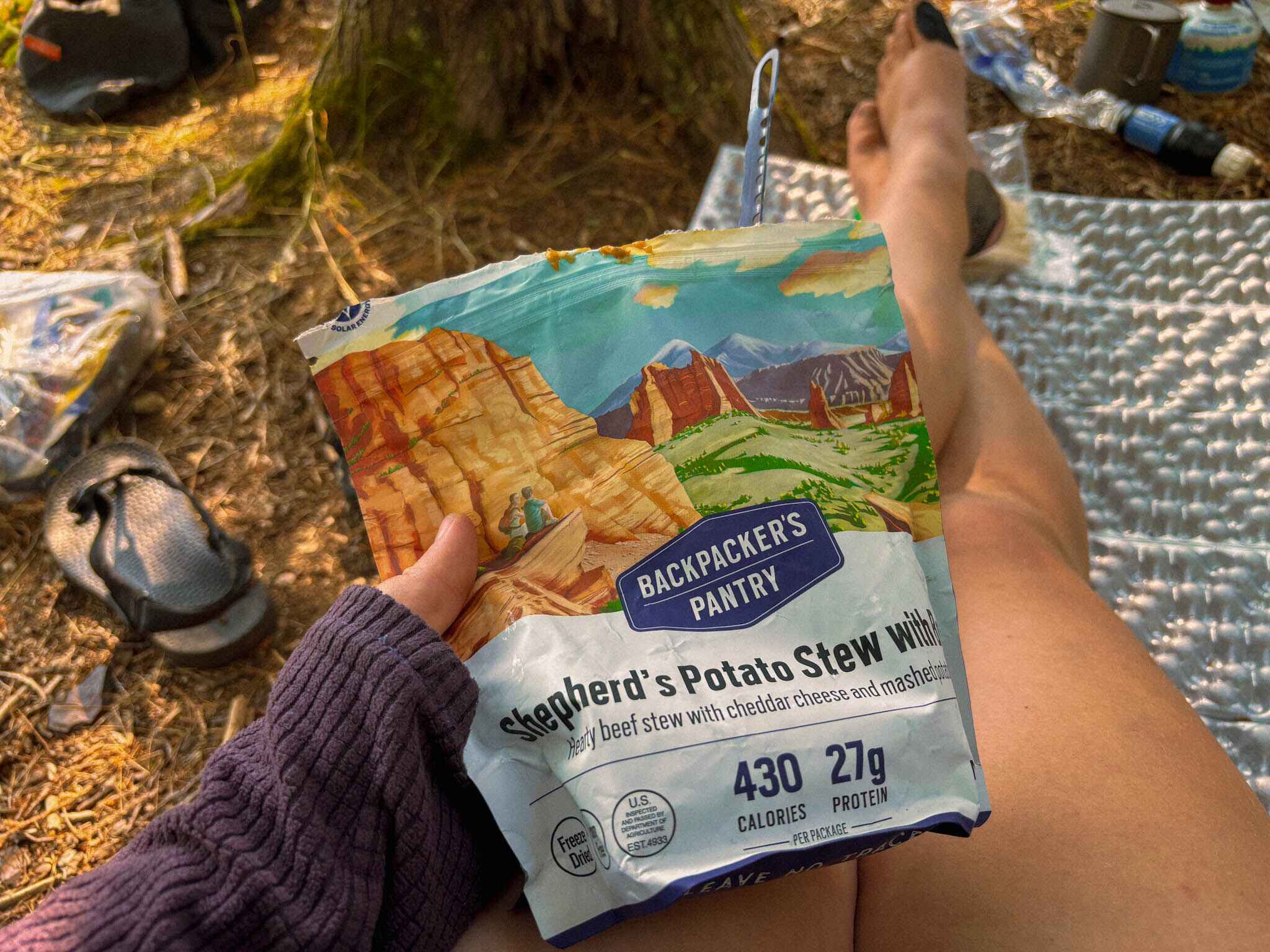 A hiker holds Shepherds potato stew to the camera while lounging on a sleeping pad