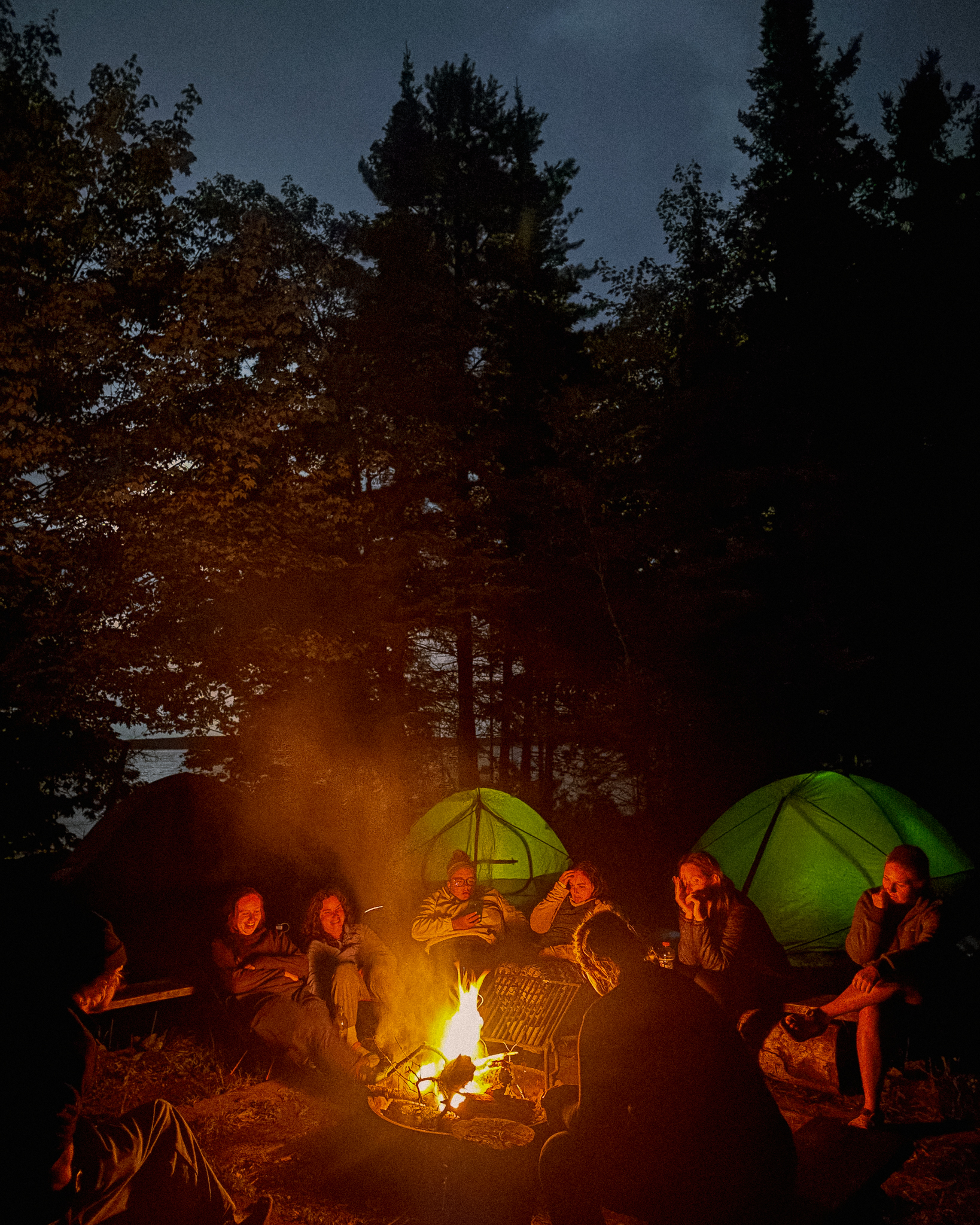 Image of campers gathered around an evening campfire with tents glowing in the background