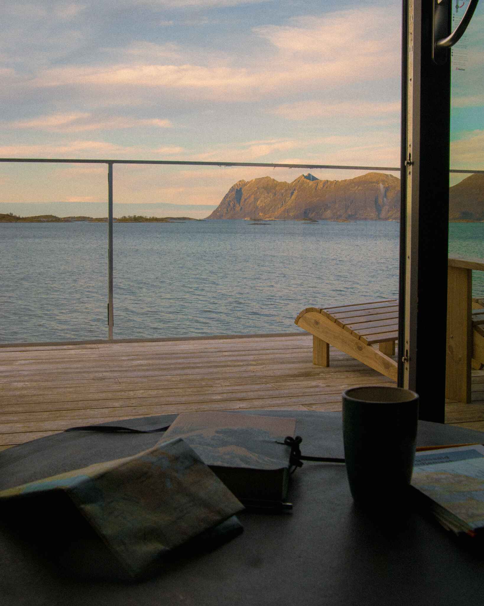 Image from an Aurora suite at Hamn i Senja; a journal, maps and a cup of coffee sit on a table with a backdrop of a balcony looking out over the ocean with mountains in the background