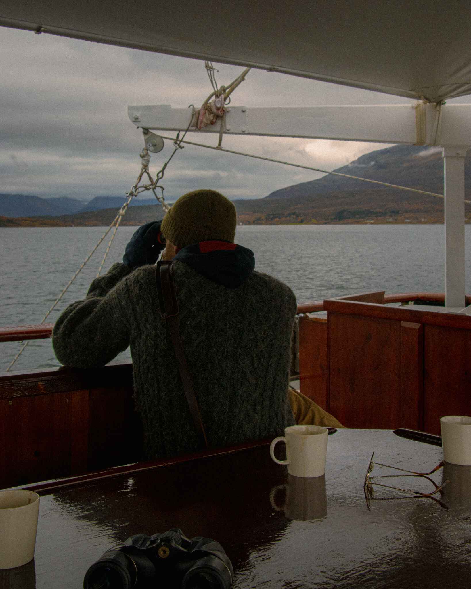 image of a man in a wool sweater on a boat with binoculars looking out over a Norwegian fjord near tromso, norway on a moody day